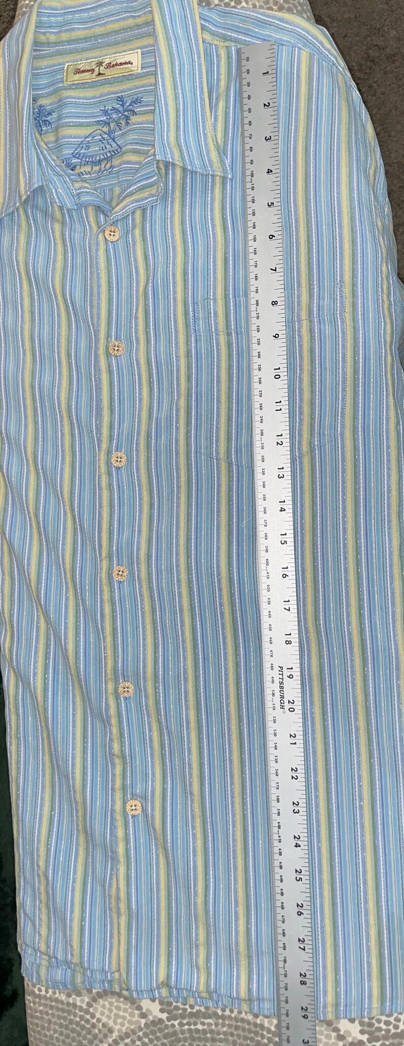 Puede incluir: Camisa abotonada con rayas verticales en azul claro, amarillo y blanco. La camisa tiene un bolsillo en el pecho y mide aproximadamente 76 cm de largo. La marca "Tommy Bahama" es visible en el cuello.