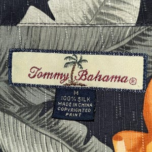 Puede incluir: Primer plano de la etiqueta de una camisa de seda Tommy Bahama. La etiqueta rectangular tiene un borde azul y el nombre de la marca en escritura roja. La camisa tiene un estampado de hojas tropicales en tonos grises, negros y naranjas. La etiqueta dice "100% Silk, Made in China, Copyrighted Print."