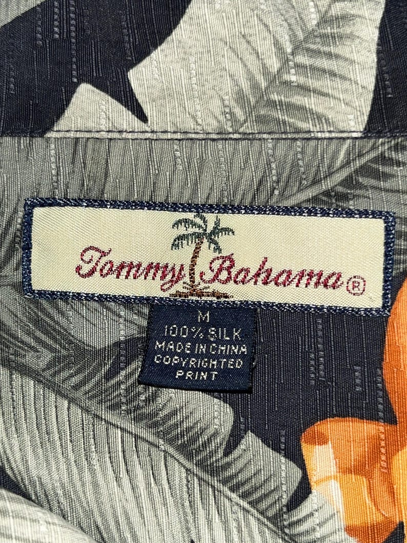 Puede incluir: Primer plano de la etiqueta de una camisa de seda Tommy Bahama. La etiqueta rectangular tiene un borde azul y el nombre de la marca en escritura roja. La camisa tiene un estampado de hojas tropicales en tonos grises, negros y naranjas. La etiqueta dice "100% Silk, Made in China, Copyrighted Print."