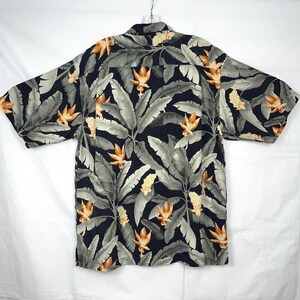 Puede incluir: Una camisa de manga corta con un estampado floral tropical. La camisa presenta un fondo azul marino oscuro con hojas grises grandes y flores naranjas y amarillas. Cuelga de una percha sobre un fondo blanco.