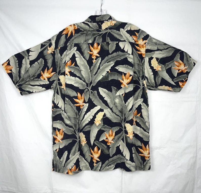 Puede incluir: Una camisa de manga corta con un estampado floral tropical. La camisa presenta un fondo azul marino oscuro con hojas grises grandes y flores naranjas y amarillas. Cuelga de una percha sobre un fondo blanco.
