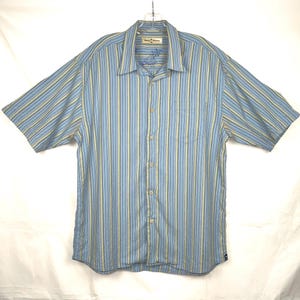 Puede incluir: Camisa de manga corta a rayas azules y amarillas. La camisa tiene una botonadura frontal, un bolsillo en el pecho y un cuello. La marca "Tommy Bahama" es visible en el cuello.