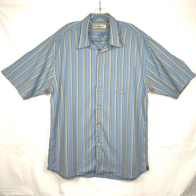 Puede incluir: Camisa de manga corta a rayas azules y amarillas. La camisa tiene una botonadura frontal, un bolsillo en el pecho y un cuello. La marca "Tommy Bahama" es visible en el cuello.