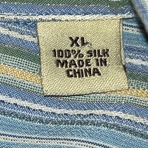 Puede incluir: Primer plano de una etiqueta de tela con el texto "XL 100% SILK MADE IN CHINA". La etiqueta es beige y est&aacute; cosida a una tela a rayas azules, verdes y amarillas. La tela parece ser una mezcla de seda.