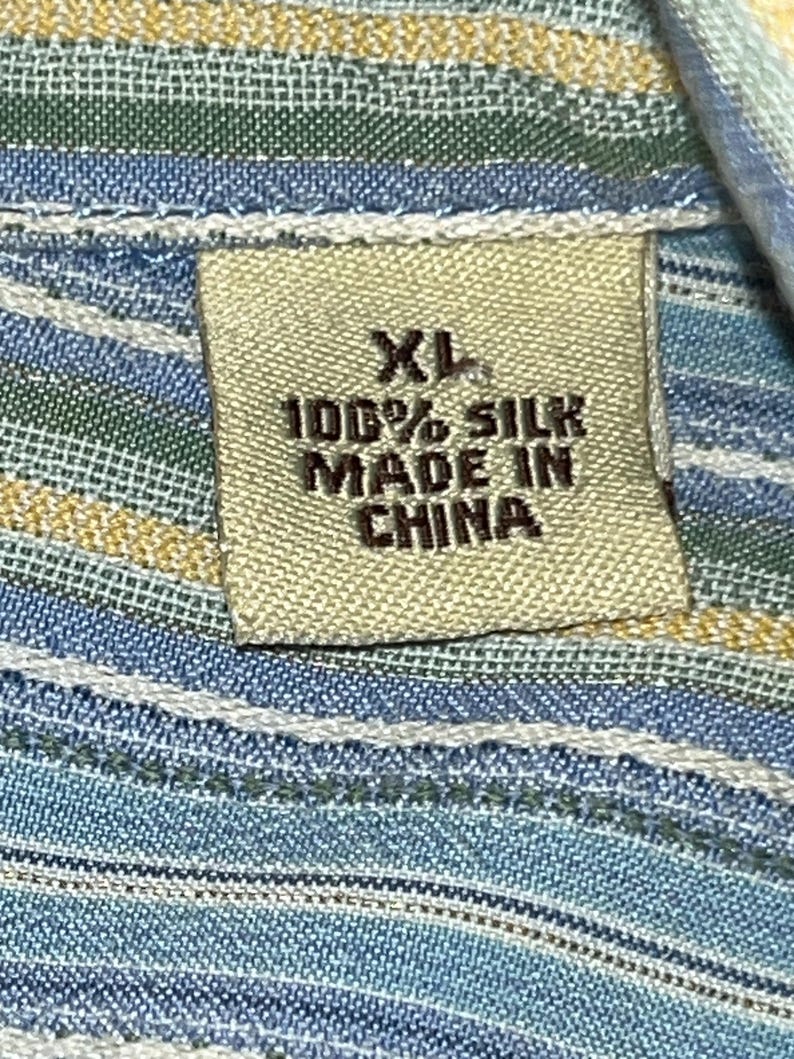 Puede incluir: Primer plano de una etiqueta de tela con el texto "XL 100% SILK MADE IN CHINA". La etiqueta es beige y est&aacute; cosida a una tela a rayas azules, verdes y amarillas. La tela parece ser una mezcla de seda.