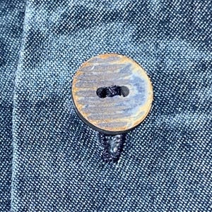 Może przedstawiać: Zbliżenie okrągłego, drewnianego guzika w niebiesko-brązowej kolorystyce, przyszytego do ciemnoniebieskiego dżinsu. Guzik ma dwa otwory do szycia i teksturowaną powierzchnię. Materiał dżinsowy ma subtelny, tkany wz&oacute;r.