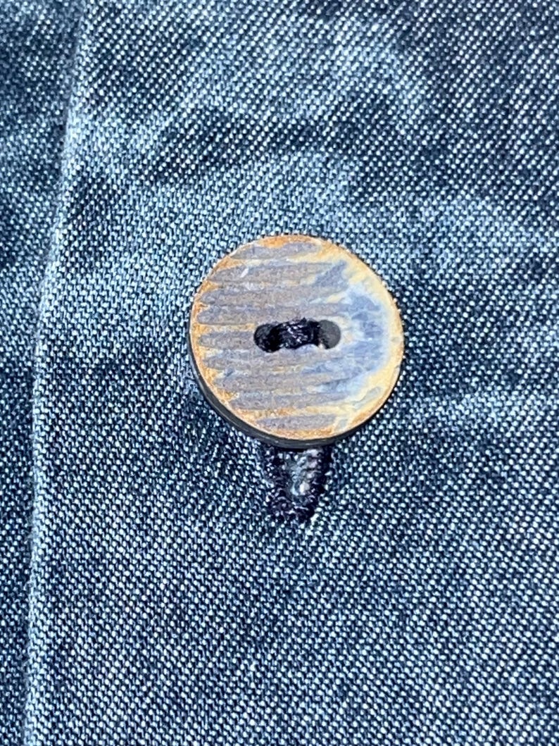 Może przedstawiać: Zbliżenie okrągłego, drewnianego guzika w niebiesko-brązowej kolorystyce, przyszytego do ciemnoniebieskiego dżinsu. Guzik ma dwa otwory do szycia i teksturowaną powierzchnię. Materiał dżinsowy ma subtelny, tkany wz&oacute;r.