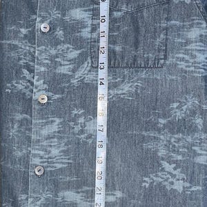 Może przedstawiać: Niebieska koszula zapinana na guziki z tropikalnym nadrukiem. Koszula ma kieszeń na piersi i widoczna jest nazwa marki "Tommy Bahama". Taśma miernicza pokazuje, że koszula ma około 79 cm długości.