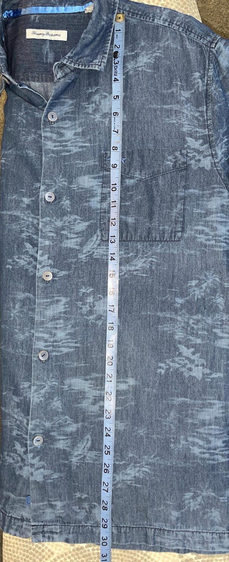Może przedstawiać: Niebieska koszula zapinana na guziki z tropikalnym nadrukiem. Koszula ma kieszeń na piersi i widoczna jest nazwa marki "Tommy Bahama". Taśma miernicza pokazuje, że koszula ma około 79 cm długości.