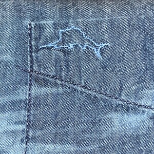 Może przedstawiać: Zbliżenie kieszeni niebieskiej koszuli dżinsowej z jasno niebieskim haftowanym konturem wyspy. Kieszeń jest przeszyta ciemnoniebieską nicią, a koszula ma teksturowany wygląd.