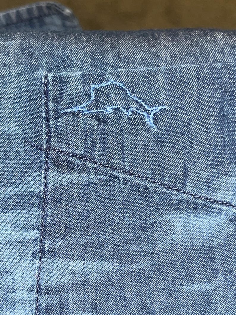 Może przedstawiać: Zbliżenie kieszeni niebieskiej koszuli dżinsowej z jasno niebieskim haftowanym konturem wyspy. Kieszeń jest przeszyta ciemnoniebieską nicią, a koszula ma teksturowany wygląd.