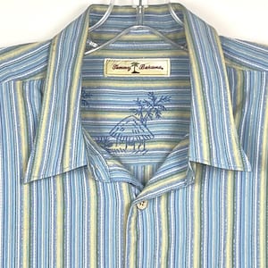 Puede incluir: Camisa a rayas verticales en tonos azul, amarillo y blanco. Presenta cuello, bolsillo en el pecho y botones de madera. La etiqueta dice "Tommy Bahama" con un logotipo de palmera y un dise&ntilde;o tropical bordado.