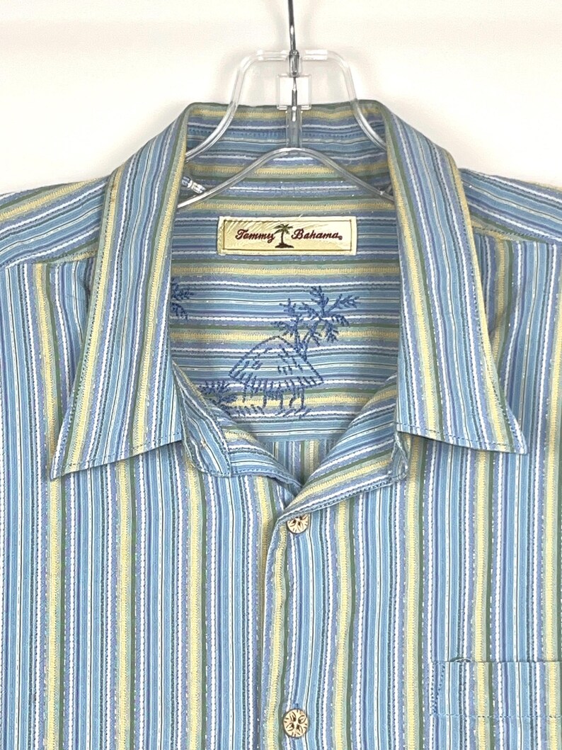 Puede incluir: Camisa a rayas verticales en tonos azul, amarillo y blanco. Presenta cuello, bolsillo en el pecho y botones de madera. La etiqueta dice "Tommy Bahama" con un logotipo de palmera y un dise&ntilde;o tropical bordado.