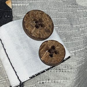 Puede incluir: Primer plano de dos botones redondos marrones con un dise&ntilde;o en forma de cruz en el centro. Los botones est&aacute;n hechos de un material natural y est&aacute;n adheridos a una tela blanca. El fondo es una tela gris claro texturizada.