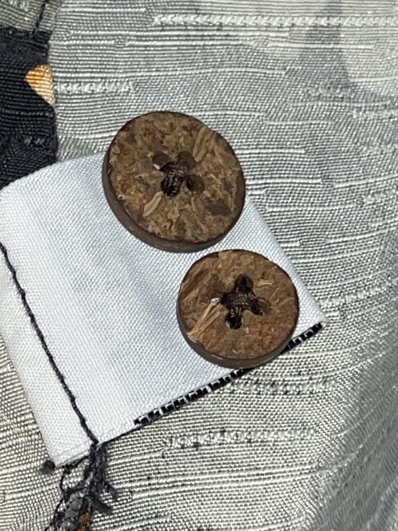 Puede incluir: Primer plano de dos botones redondos marrones con un dise&ntilde;o en forma de cruz en el centro. Los botones est&aacute;n hechos de un material natural y est&aacute;n adheridos a una tela blanca. El fondo es una tela gris claro texturizada.