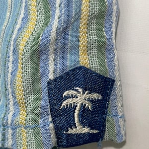 Puede incluir: Primer plano de una tela a rayas azules y blancas con detalles amarillos y verdes. Un parche azul marino con un dise&ntilde;o de palmera bordada en blanco est&aacute; cosido a la tela. La tela tiene una apariencia texturizada.