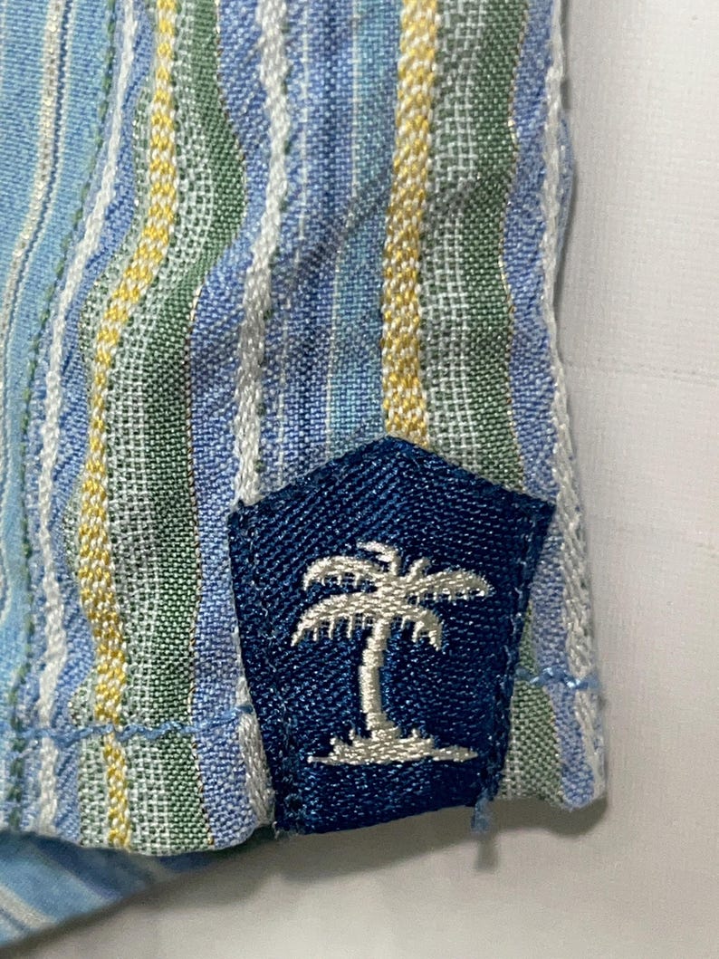 Puede incluir: Primer plano de una tela a rayas azules y blancas con detalles amarillos y verdes. Un parche azul marino con un dise&ntilde;o de palmera bordada en blanco est&aacute; cosido a la tela. La tela tiene una apariencia texturizada.