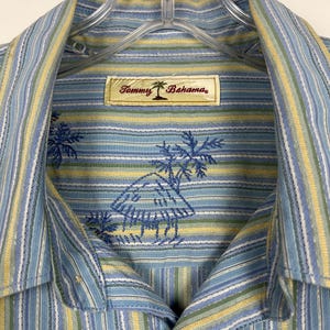 Puede incluir: Camisa a rayas azules y amarillas con un logotipo de palmera. La camisa presenta un patr&oacute;n de rayas azul claro y amarillo con palmeras bordadas y la etiqueta de la marca "Tommy Bahama".