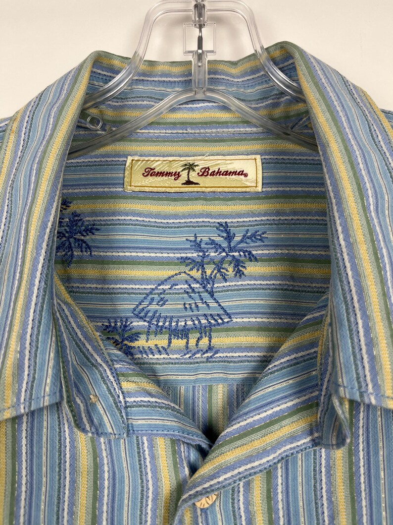 Puede incluir: Camisa a rayas azules y amarillas con un logotipo de palmera. La camisa presenta un patr&oacute;n de rayas azul claro y amarillo con palmeras bordadas y la etiqueta de la marca "Tommy Bahama".