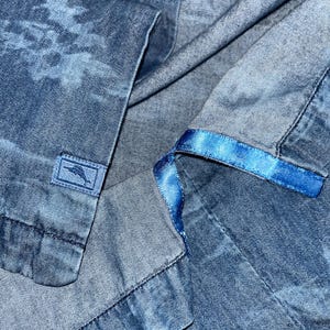 Może przedstawiać: Zbliżenie niebieskiej koszuli dżinsowej z subtelnym wzorem przypominającym chmury. Koszula posiada małą prostokątną metkę z logo oraz niebieską satynową wstążkę wzdłuż wewnętrznego kołnierza.