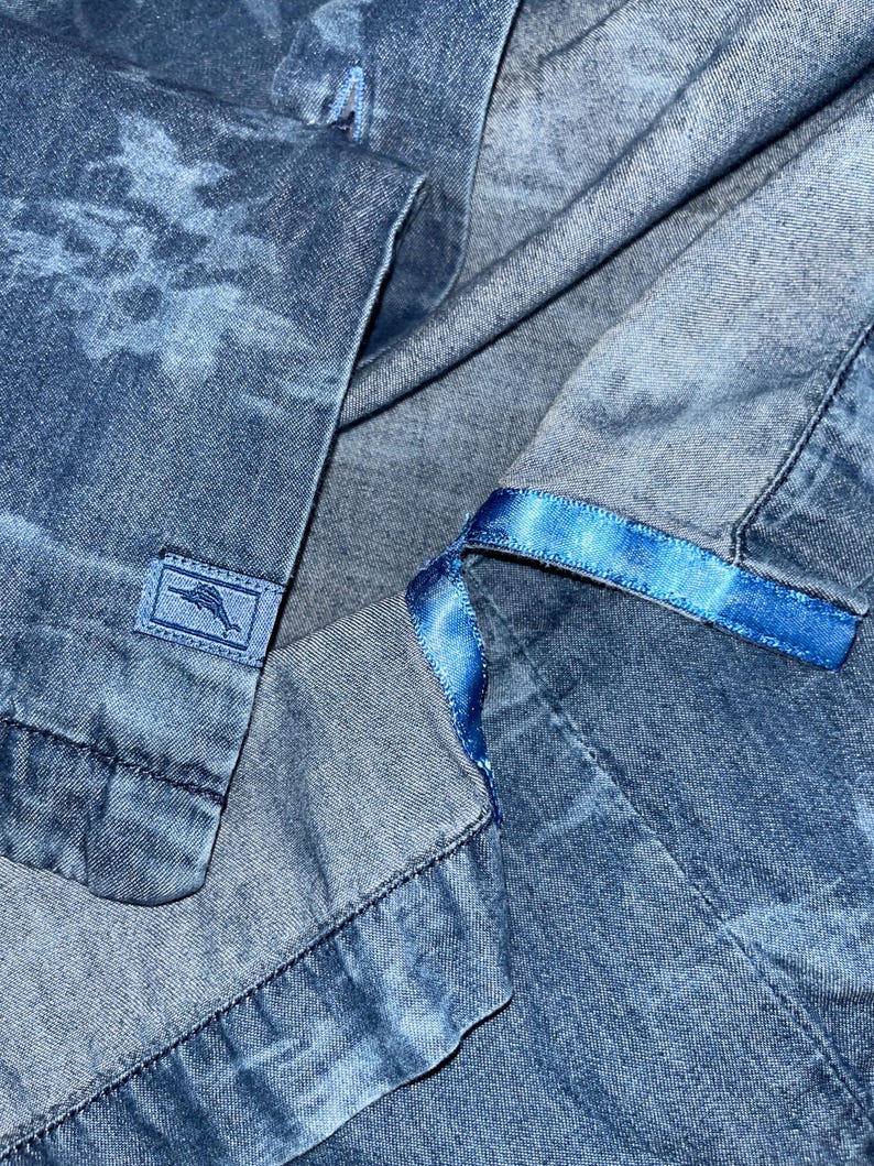 Może przedstawiać: Zbliżenie niebieskiej koszuli dżinsowej z subtelnym wzorem przypominającym chmury. Koszula posiada małą prostokątną metkę z logo oraz niebieską satynową wstążkę wzdłuż wewnętrznego kołnierza.