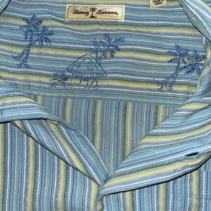 Puede incluir: Camisa abotonada a rayas azules, amarillas y blancas con palmeras bordadas y una etiqueta Tommy Bahama. La camisa es de seda y talla XL.
