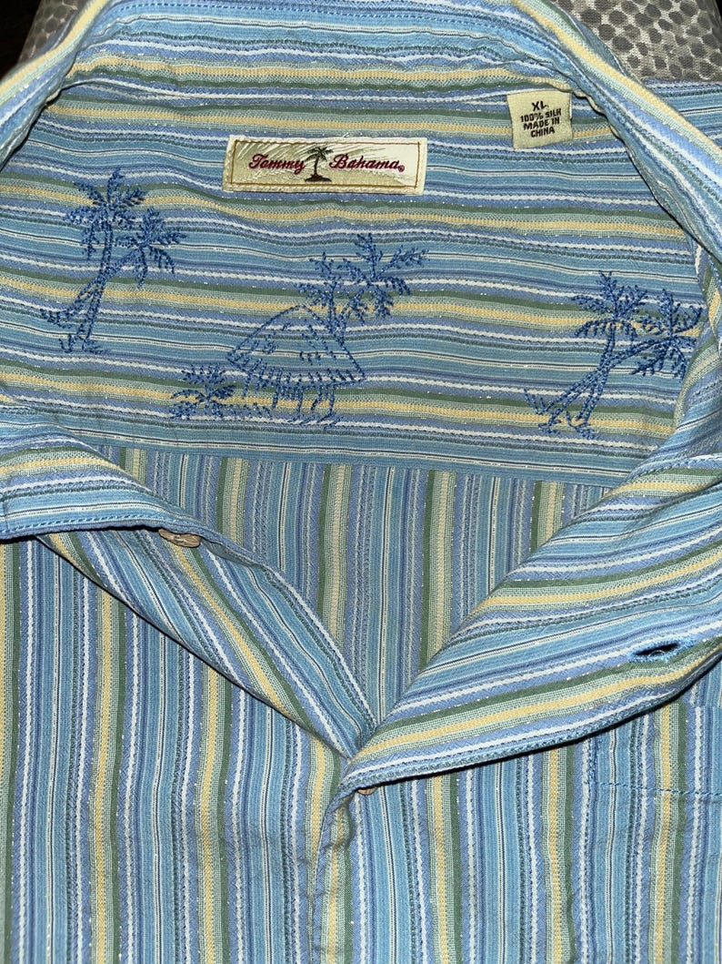 Puede incluir: Camisa abotonada a rayas azules, amarillas y blancas con palmeras bordadas y una etiqueta Tommy Bahama. La camisa es de seda y talla XL.