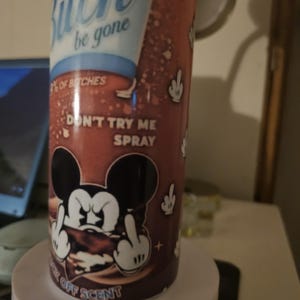 Puede incluir: Un frasco de spray cil&iacute;ndrico con un fondo marr&oacute;n y texto blanco que dice "Bitch be gone". La botella presenta un personaje de rat&oacute;n de dibujos animados haciendo una peineta. Tambi&eacute;n es visible el texto "Don't try me spray".