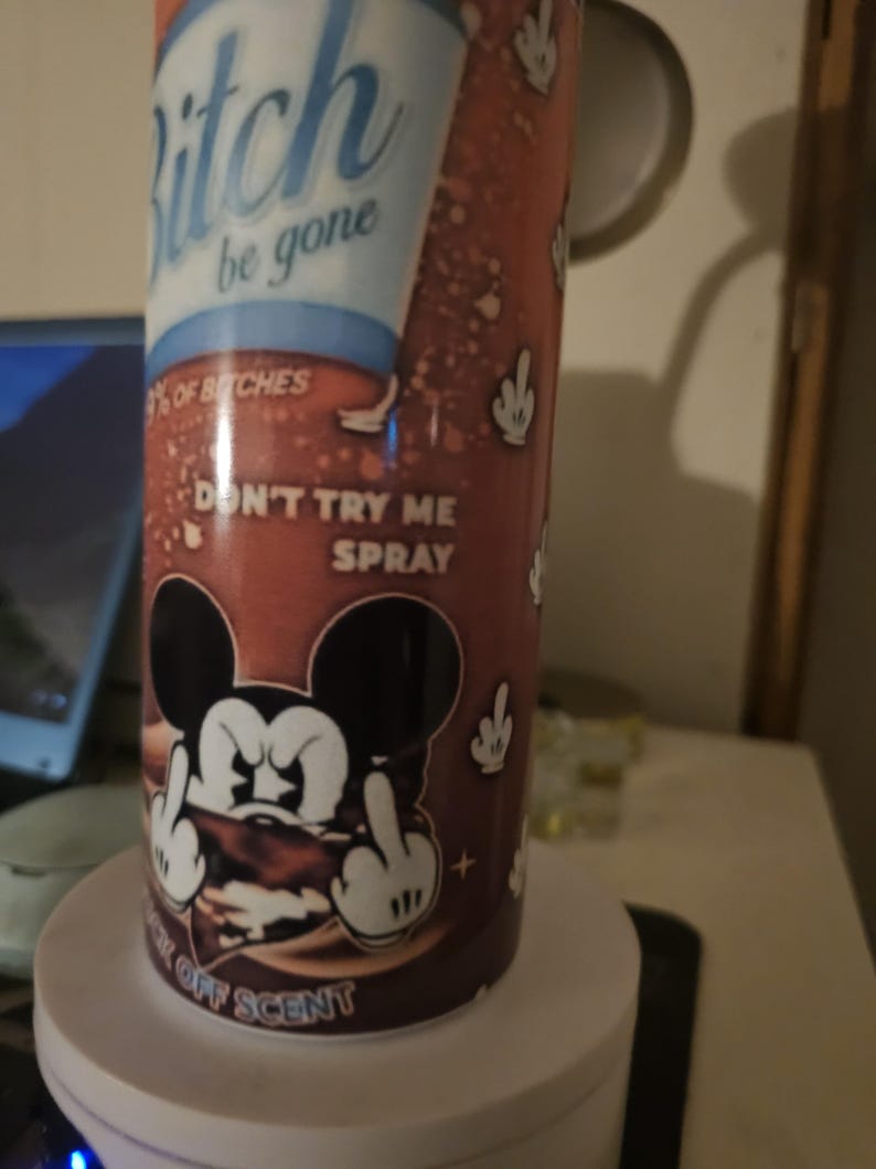 Puede incluir: Un frasco de spray cil&iacute;ndrico con un fondo marr&oacute;n y texto blanco que dice "Bitch be gone". La botella presenta un personaje de rat&oacute;n de dibujos animados haciendo una peineta. Tambi&eacute;n es visible el texto "Don't try me spray".