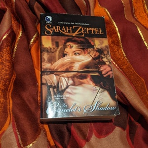 May include: A book cover with a woman in a hooded cloak holding a bow and arrow. The title of the book is "In Camelot's Shadow" by Sarah Zettel. The book cover is a light brown color with a gold border.
