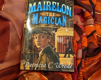 May include: A paperback book titled "Mairelon the Magician" by Patricia C. Wrede. The cover features a young person with short brown hair wearing a cap and a dark jacket. The background is a city street with a building and a sign that says "Is it possible? Could his magic be...real?"