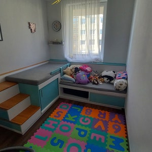 Könnte beinhalten: Ein hellblaues und weißes Holzbett mit einer grauen Matratze und einer bunten Alphabet-Schaumstoffmatte auf dem Boden. Das Bett hat eine eingebaute Treppe und Schubladen.