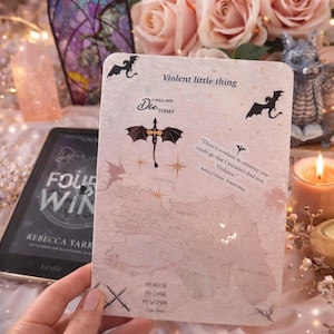 Puede incluir: Una tarjeta con el texto "Violent little thing" y una ilustración de dragón, con las palabras "I will not die today". La tarjeta se sostiene cerca de un Kindle y está rodeada de rosas y velas. La tarjeta también tiene el texto "My house, my chair, my woman".