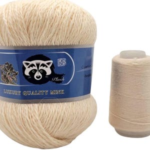 May include: Two skeins of cream-coloured yarn. One is a large ball with a blue label featuring a raccoon illustration and the text "Luxury Quality Mink". The other is a smaller cone of yarn.