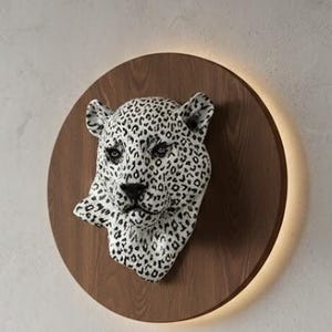 May include: A wall-mounted leopard head sculpture with black spots on a white base, set against a dark wood circular panel. The panel is illuminated by a warm light around its edge, creating a striking contrast and a modern aesthetic.