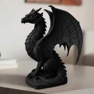 Può includere: Una statuetta di drago nero con squame dettagliate e grandi ali. Il drago è seduto su una base rettangolare. La statua è esposta su un tavolo colorato chiaro con una cornice in metallo nero, in un salotto.