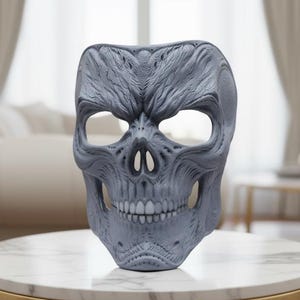 May include: A gray skull mask with detailed facial features, including eye sockets, a nose cavity, and teeth. The mask is displayed on a marble-topped table with gold legs, set against a blurred interior background.