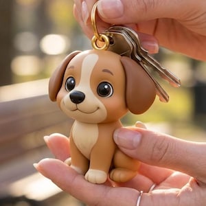 Pode incluir: Um chaveiro de cachorro marrom e branco com um chaveiro dourado e chaves. O cachorro tem olhos grandes e uma expressão sorridente. O chaveiro é segurado nas mãos de uma pessoa.