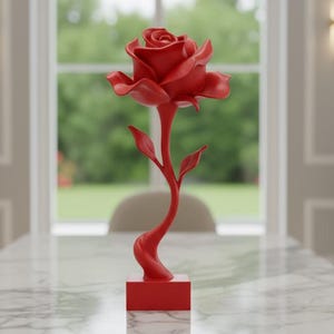 Puede incluir: Una vibrante escultura de rosa roja, con una flor y un tallo detallados, se exhibe sobre una mesa con tapa de mármol. La escultura se encuentra sobre un fondo de ventana e interior de tonos neutros, creando un contraste llamativo.