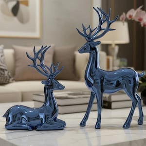 Può includere: Due statuette decorative di cervi blu, una in piedi e l'altra sdraiata, su un tavolino da caffè in marmo bianco con gambe dorate. Lo sfondo comprende un divano, cuscini e opere d'arte incorniciate. Un accessorio decorativo per la casa.