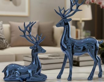 May include: Two blue decorative deer figurines. One deer is standing, the other is lying down. The figurines are placed on a white marble coffee table with gold-colored legs. The background includes a sofa, pillows, and artwork.