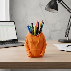 May include: An orange hoodie-shaped desk tidy filled with pens and pencils. The desk tidy is made of plastic and sits on a wooden desk. A laptop, desk lamp, and corkboard are in the background.