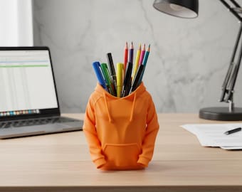 May include: An orange hoodie-shaped desk organizer filled with pens and pencils. The desk organizer is made of plastic and sits on a wooden desk. A laptop, desk lamp, and corkboard are in the background.