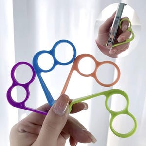 May include: Four brightly coloured phone grip holders are held in a hand. The grips are purple, blue, orange, and green. Each has two finger holes and a flat handle. An inset image shows a green grip attached to a phone.