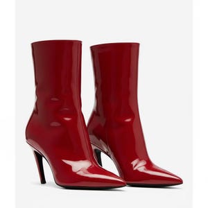Puede incluir: Un par de botines rojos brillantes, con punta y tacones de aguja. Las botas tienen un aspecto elegante y pulido y una altura media de la pantorrilla. Los tacones son negros y las botas están sobre un fondo blanco.