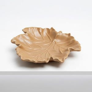 May include: A tan-colored, leaf-shaped decorative dish. The dish has detailed veining and a textured surface. The leaf dish is resting on a white surface. The dish is likely made of ceramic or a similar material.