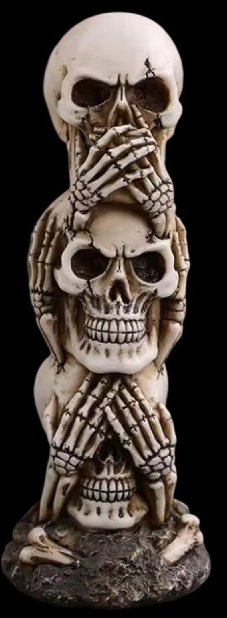 K&ouml;nnte beinhalten: Eine gestapelte Skulptur aus drei Totenk&ouml;pfen, jeder mit H&auml;nden, die Augen, Mund und Ohren bedecken. Die Sch&auml;del sind cremefarben mit detaillierten Knochenstrukturen und stehen auf einem strukturierten, dunklen Sockel. Der Hintergrund ist schwarz.