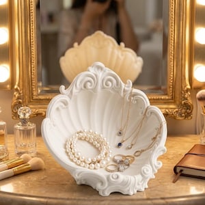 May include: A white, shell-shaped jewelry dish holds a pearl bracelet, gold necklaces, and rings. The dish sits on a marble surface, with a gold-framed mirror and makeup brushes in the background. The dish has an ornate, decorative design.