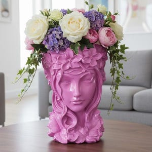 May include: A pink decorative vase shaped like a woman's face, adorned with floral details and filled with a vibrant arrangement of white roses, purple hydrangeas, and pink peonies. Green ivy spills over the sides, creating a striking display.