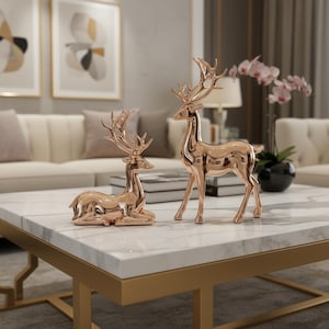 May include: Two rose gold deer figurines, one standing and one reclining, are displayed on a white marble coffee table with a gold metal frame. The deer have detailed antlers and a polished finish.